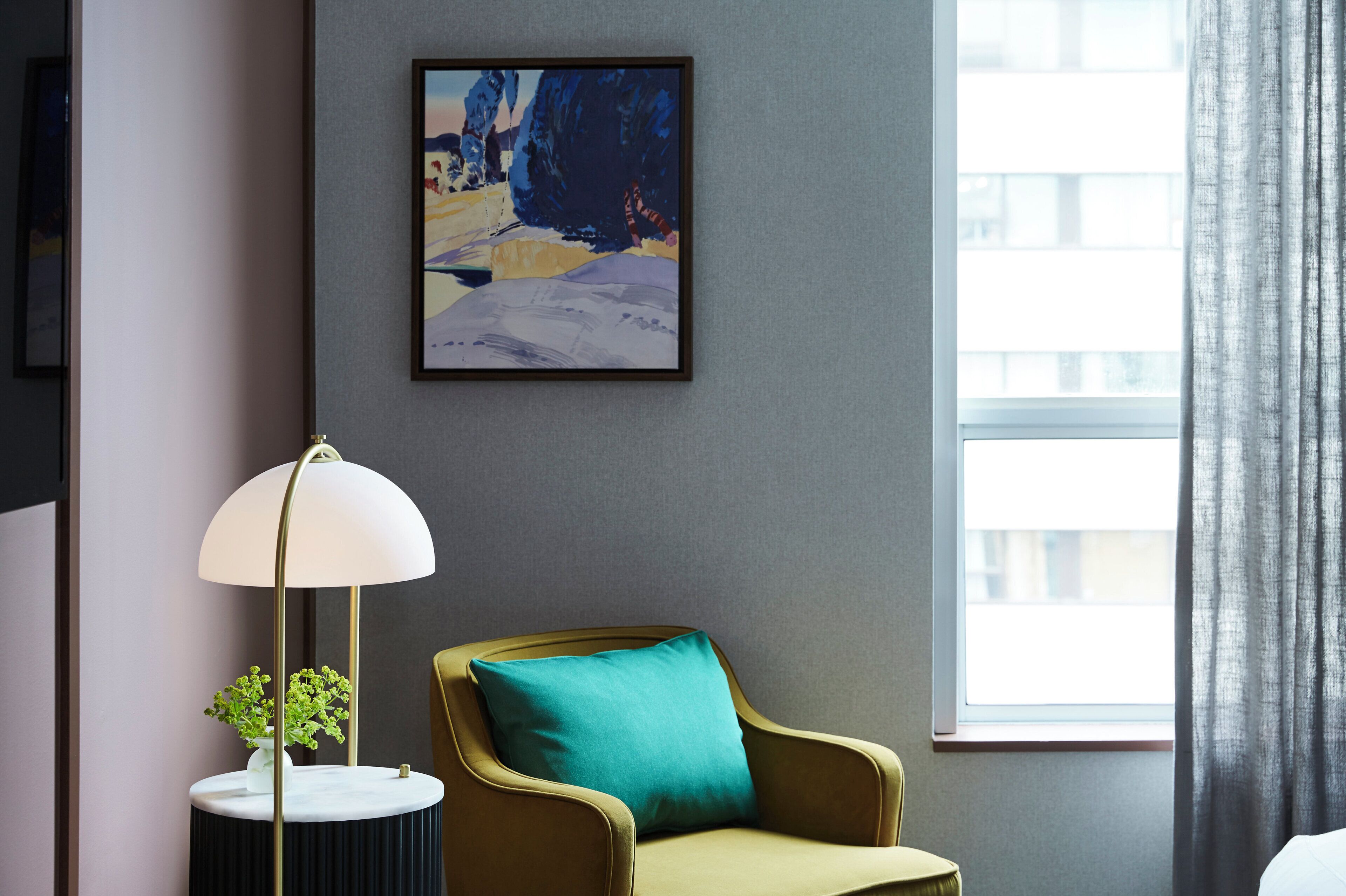 Une pièce avec un tableau encadré, une lampe, un fauteuil avec un coussin et une fenêtre avec des rideaux.