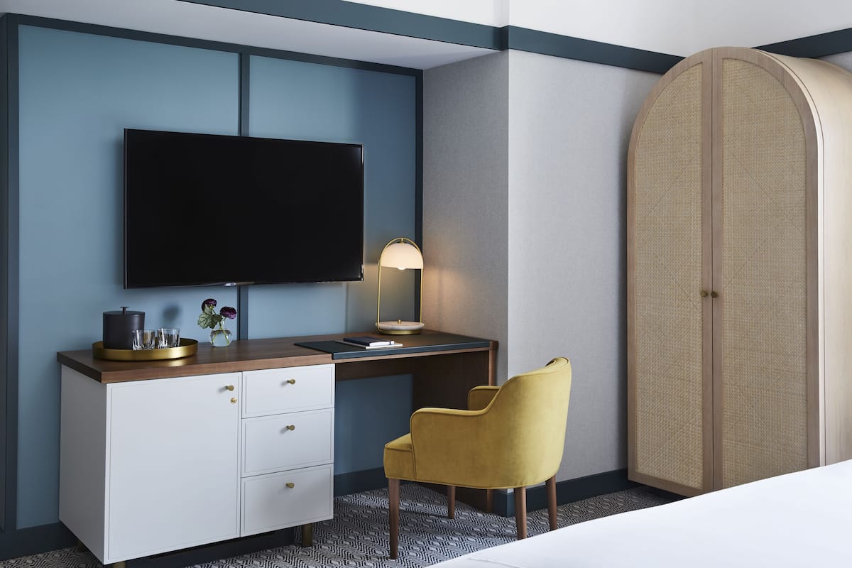 A hotel room with a bed, a desk with a flat-screen TV, a lamp, a wardrobe, and a chair.