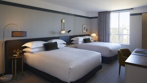 A hotel room with two beds, a desk, a chair, a lamp, and a window with a city view. - Kimpton Saint George by IHG (Toronto)