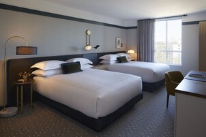 A hotel room with two beds, a desk, a chair, a lamp, and a window with a city view.