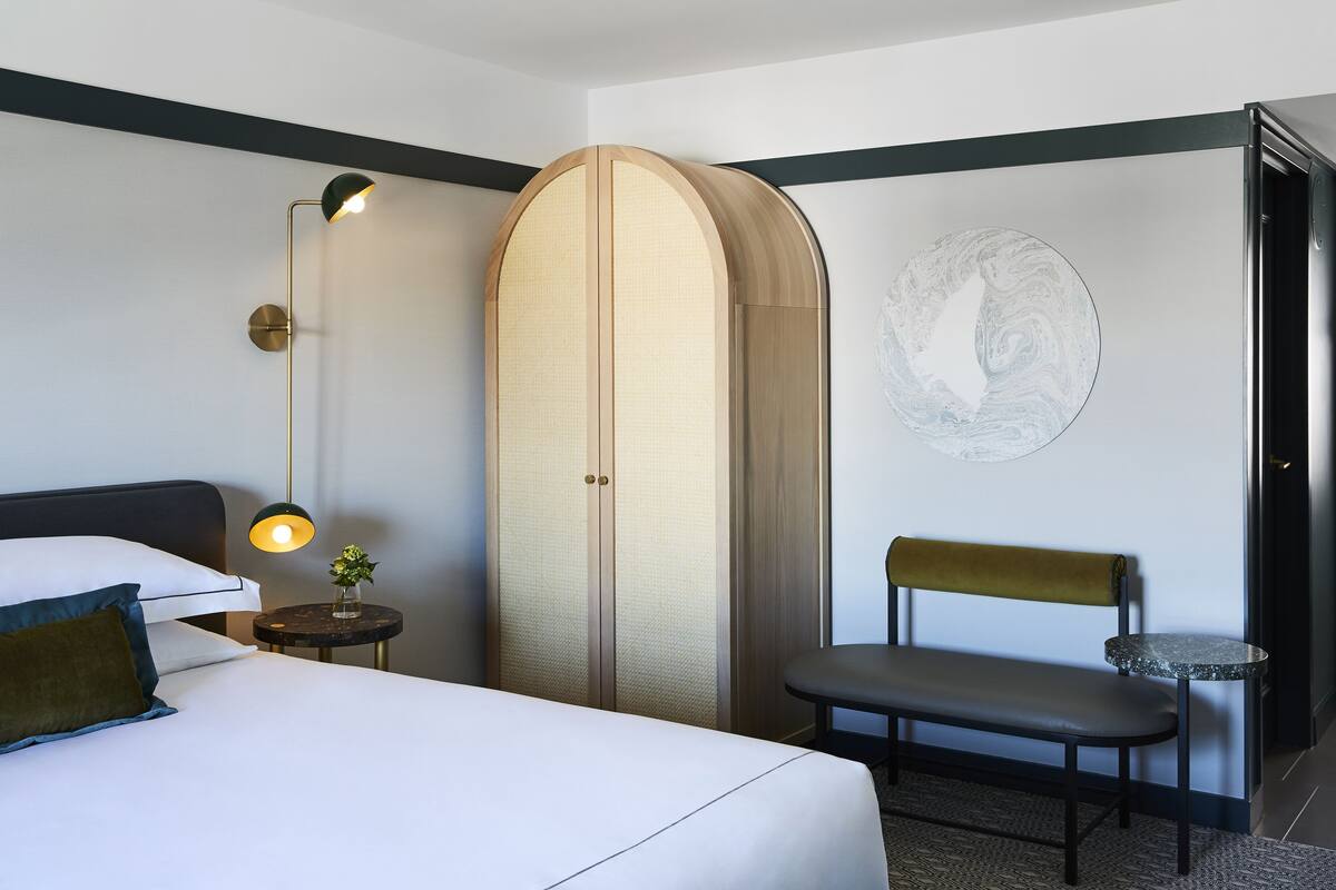 A hotel room with a bed, a bench, a small table, a lamp, and a wardrobe.