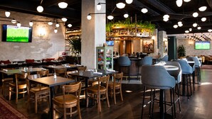 A restaurant with multiple dining tables and chairs, a bar area with shelves of bottles, and a television displaying a sports game. - Kimpton Saint George by IHG (Toronto)