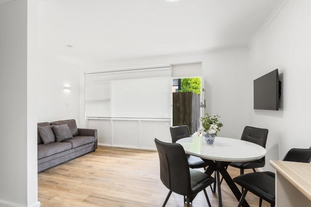 Ein modernes Zimmer mit weißer Decke, einem grauen Sofa, einem runden Tisch mit Stühlen und einem an der Wand befestigten Fernseher.