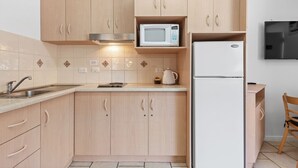 A compact kitchen with light wood cabinets, a microwave, a refrigerator, a kettle, and a sink. - Taft Motor Inn (Glenelg)