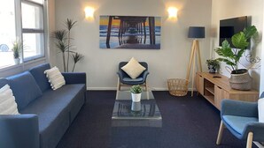 A modern living room with a blue sofa, a coffee table with a plant, a painting of a pier, and a TV on the wall. - Taft Motor Inn (Glenelg)