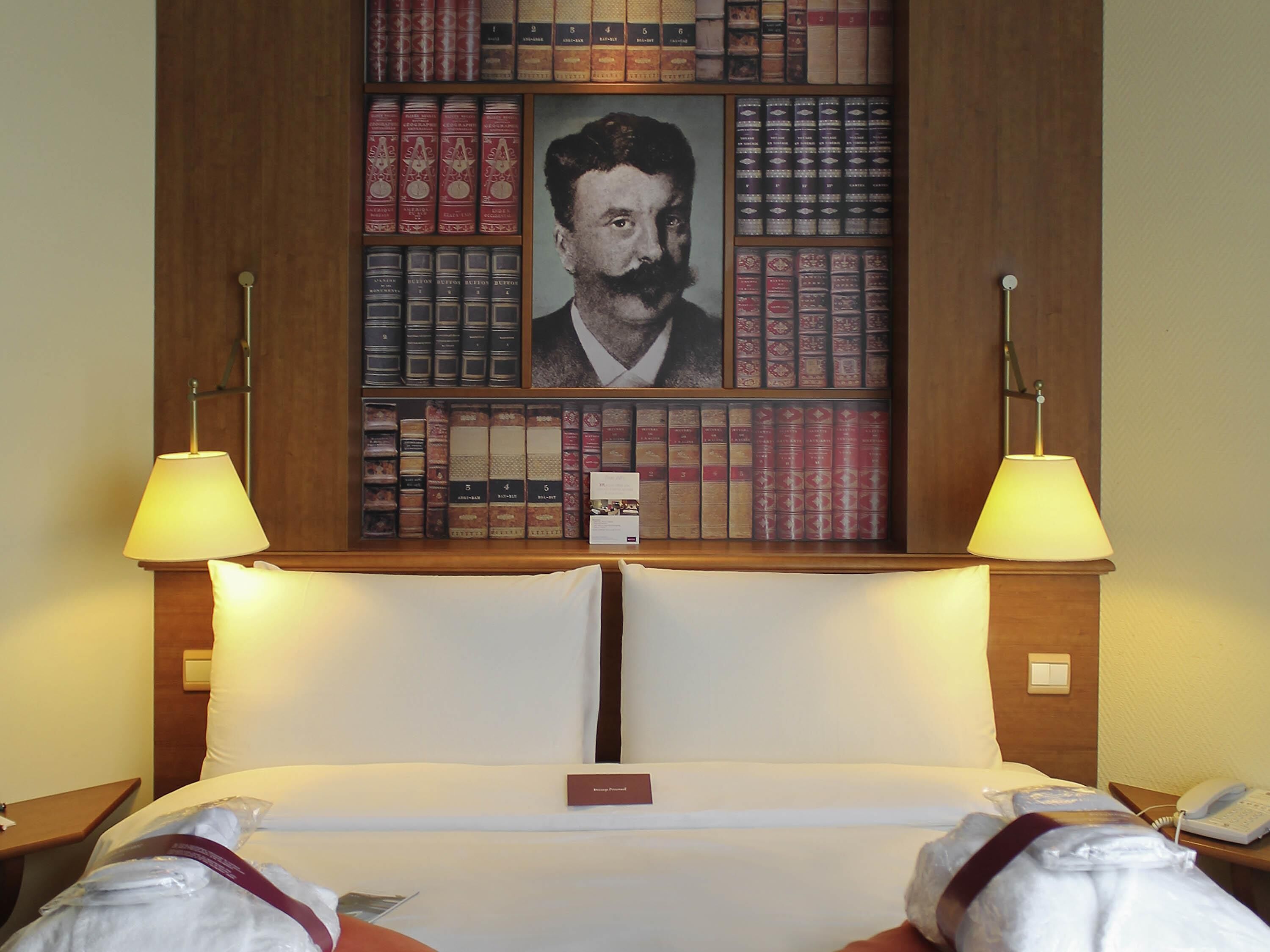 A hotel room with a large bookshelf, two bedside lamps, a bed with white linens, and a nightstand with a phone.