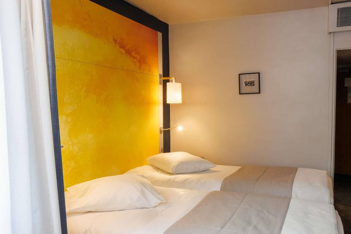 Une chambre d’hôtel avec deux lits, un mur d’accent jaune, une lampe de chevet et un tableau encadré au mur.