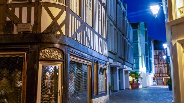 Uma estreita rua de paralelepípedos, ladeada por edifícios tradicionais em estilo enxaimel, um com uma loja à vista e outro com um letreiro indicando restaurante.