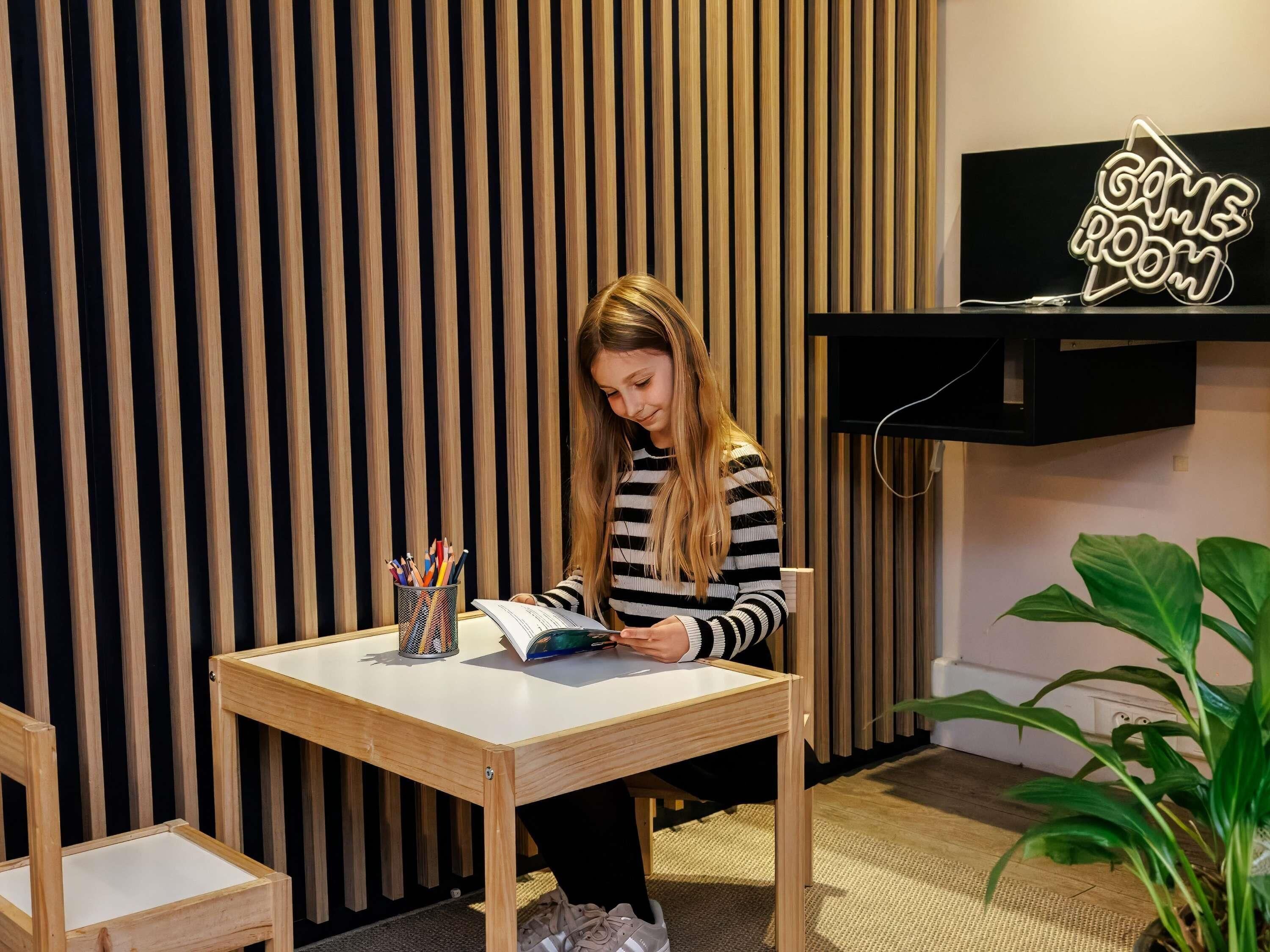 A person sitting at a wooden table with a book, a neon sign reading 'GAME ROOM' above, and a plant to the right.