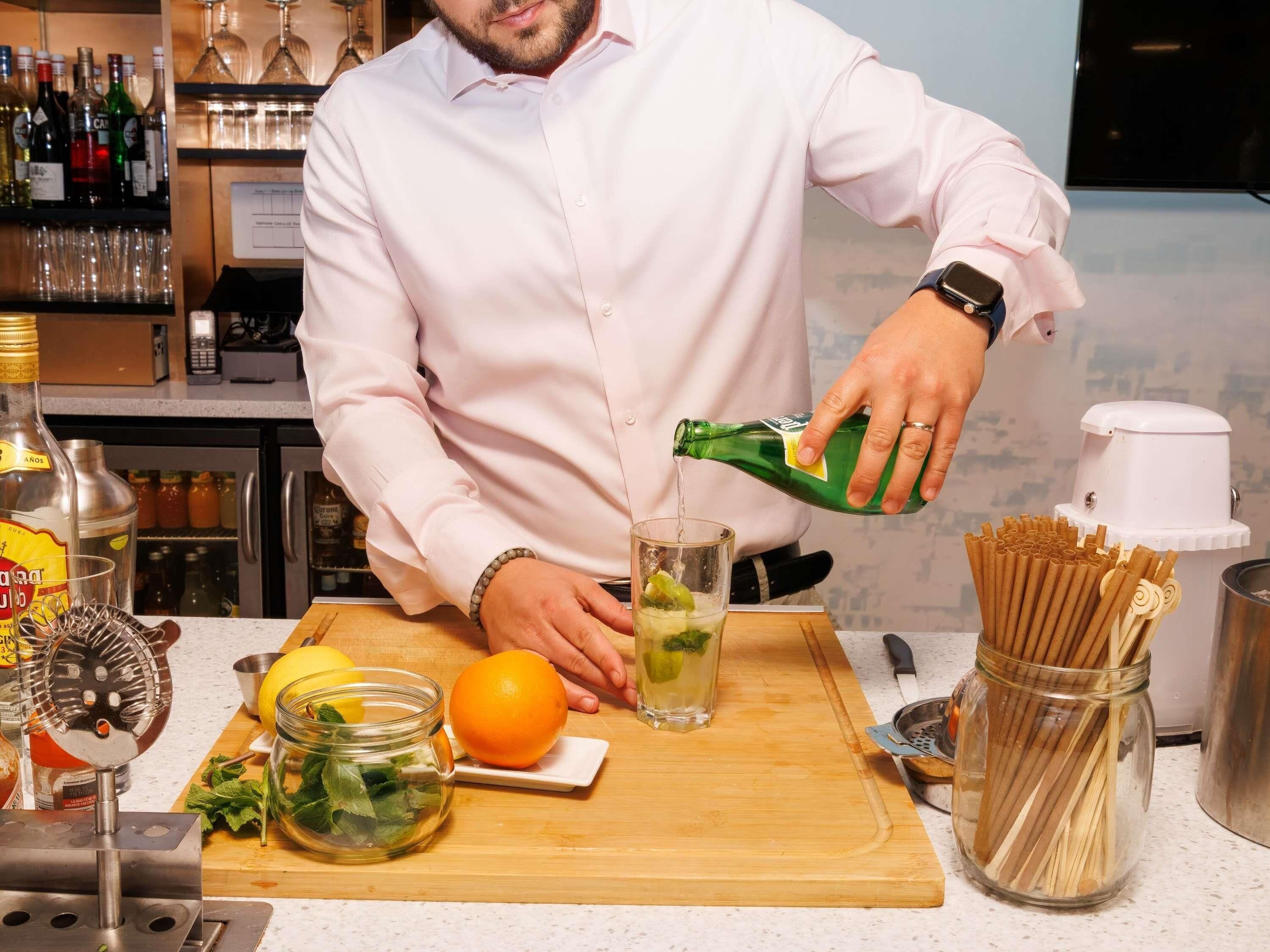 A bartender pouring a drink into a glass, with a cutting board, a jar of mint, and a lemon on the counter.
