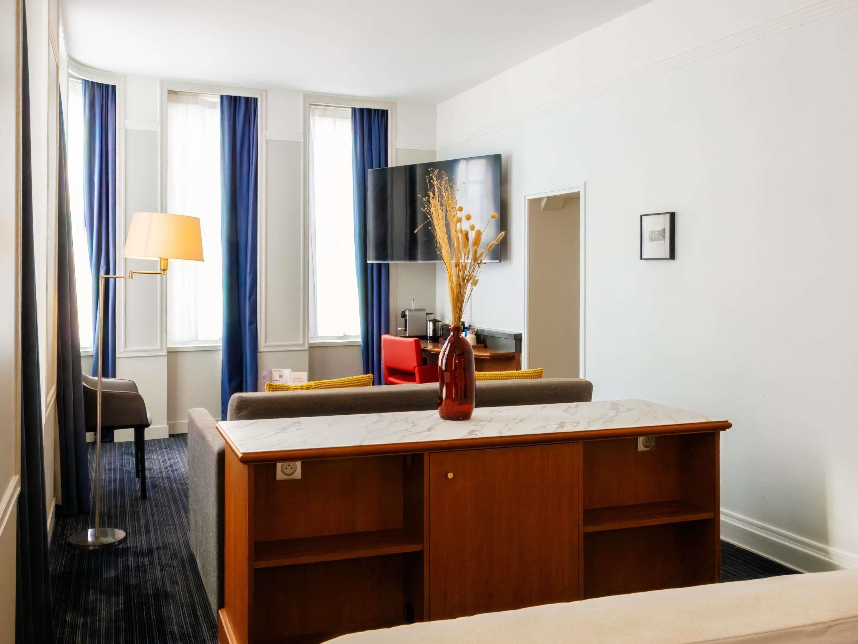 Een hotelkamer met een bed, een bureau met een vaas, een stoel, een bank, een televisie en een raam met gordijnen.