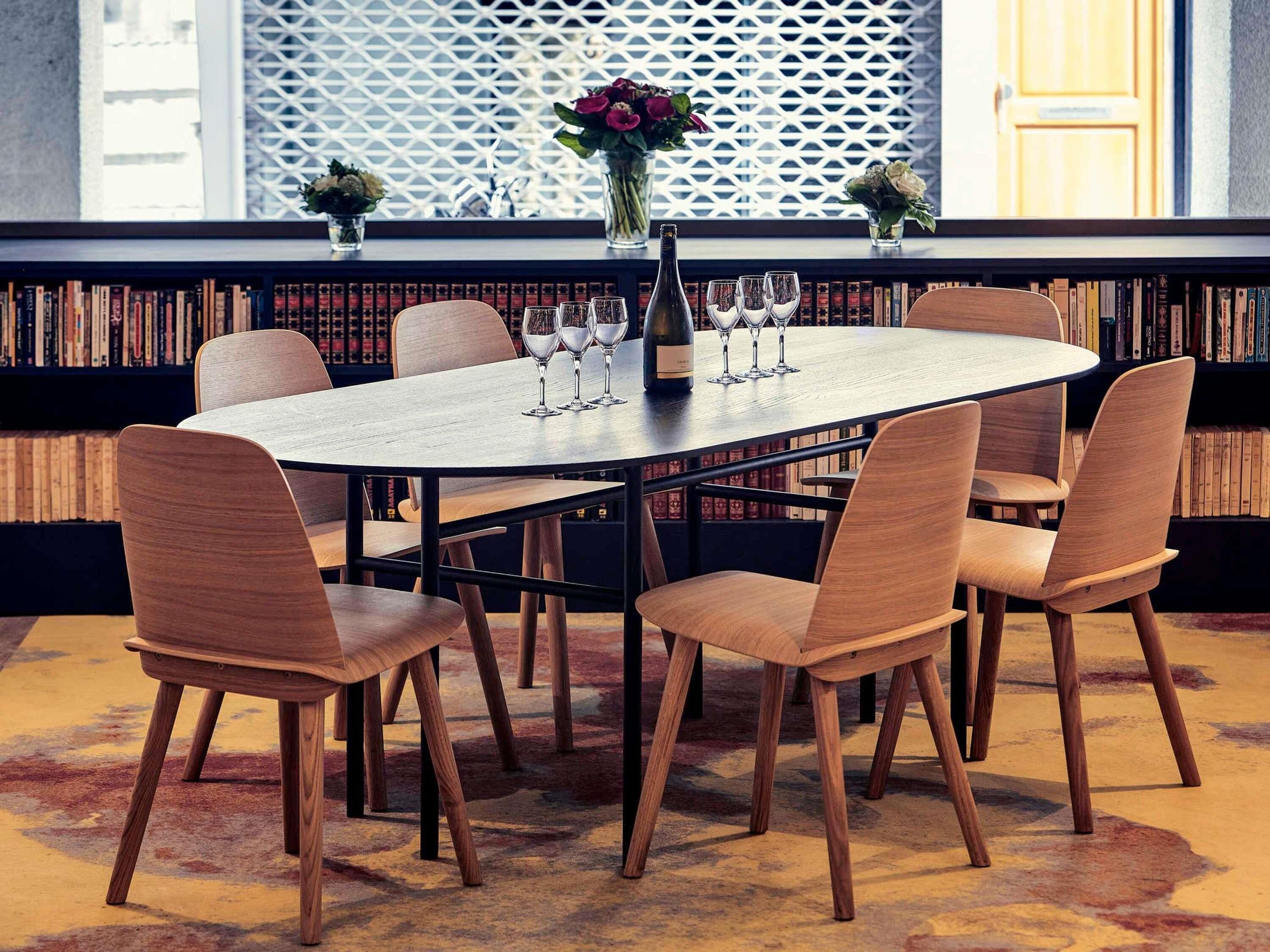 A round table with six wooden chairs, a vase with flowers, and wine glasses on a shelf with books.