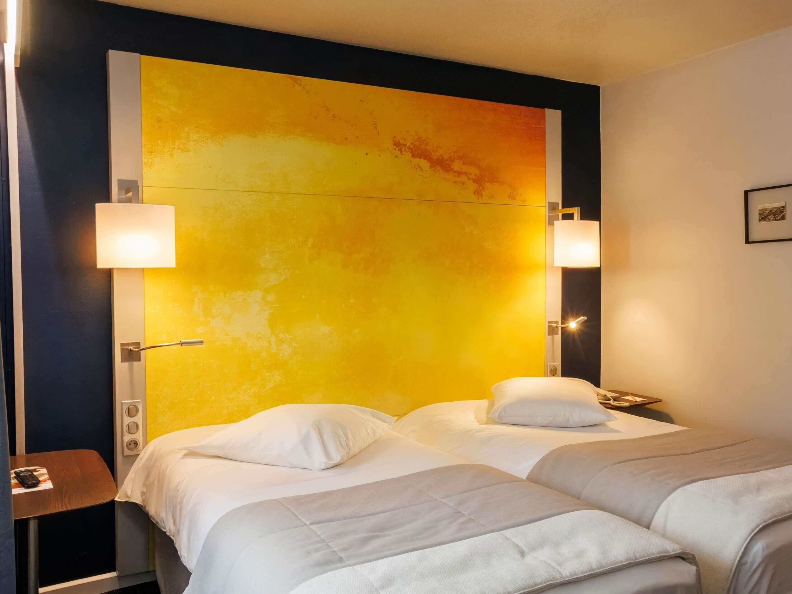 Une chambre d’hôtel avec deux lits, un grand mur jaune orné d’une œuvre abstraite et deux lampes de chevet.