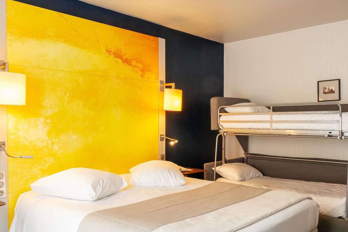 Une chambre d’hôtel avec un lit superposé et un lit simple, tous deux dotés de draps blancs. Un mur jaune vif et une section noire se trouvent sur le côté droit.