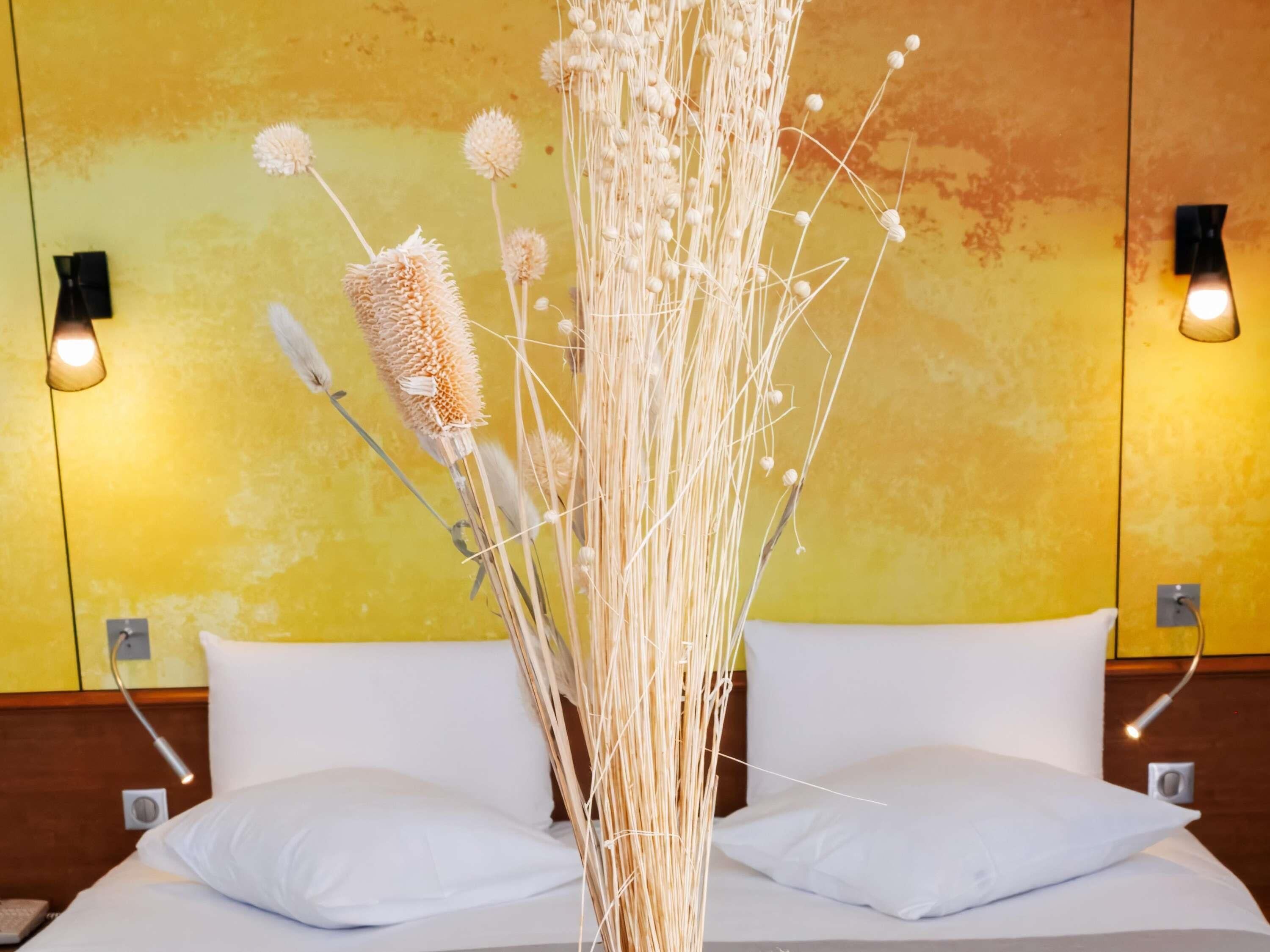 Cama com almofadas brancas, arranjo decorativo de plantas e duas luzes fixadas na parede.