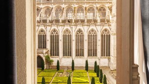 Um pátio com um jardim de layout simétrico e um edifício em estilo gótico, com grandes janelas e detalhes elaborados em pedra.