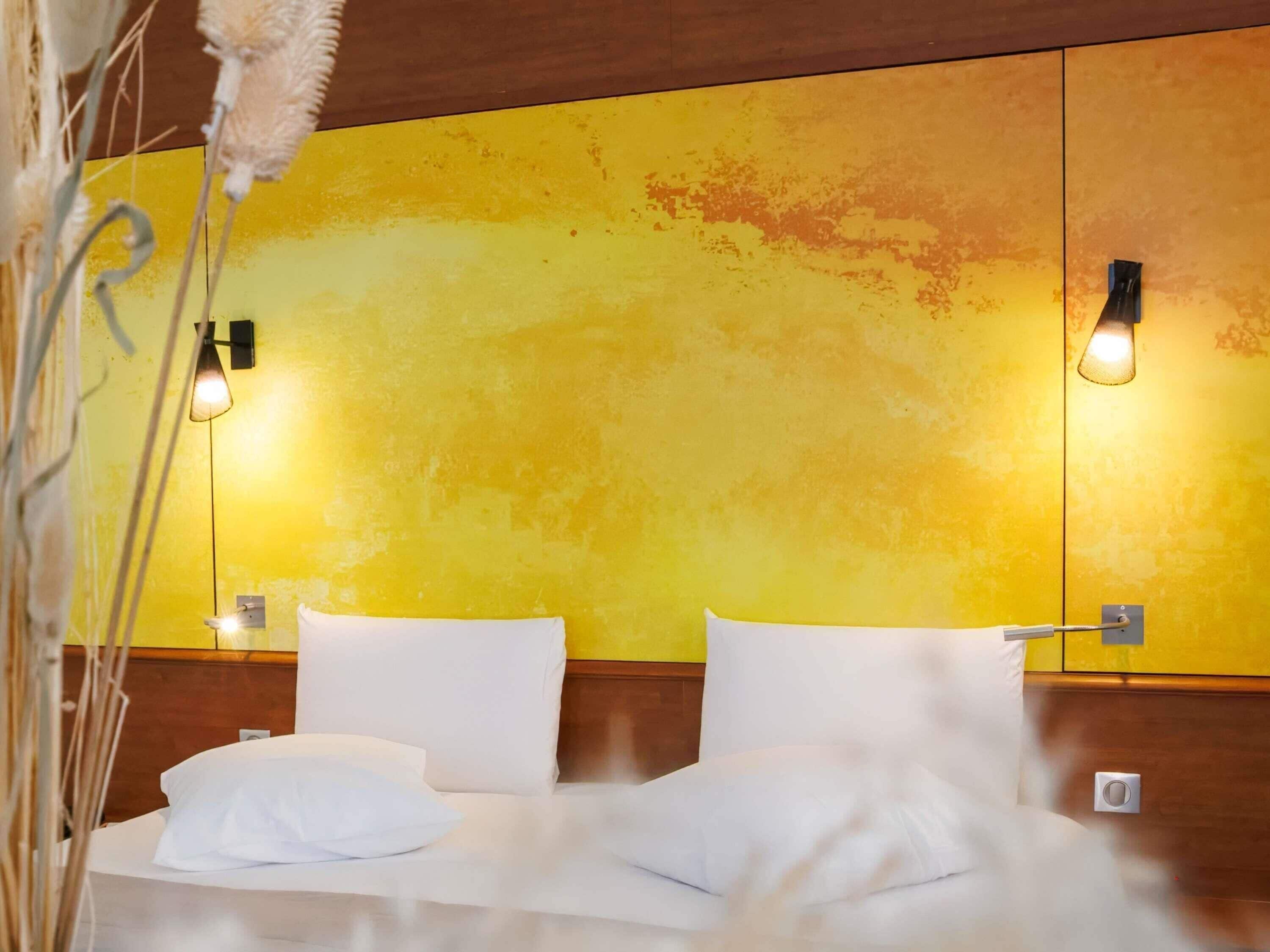 Una habitación de hotel con pared amarilla, dos lámparas de noche y una cama con ropa blanca.