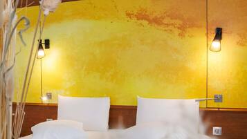 Una habitación de hotel con pared amarilla, dos lámparas de noche y una cama con ropa blanca.