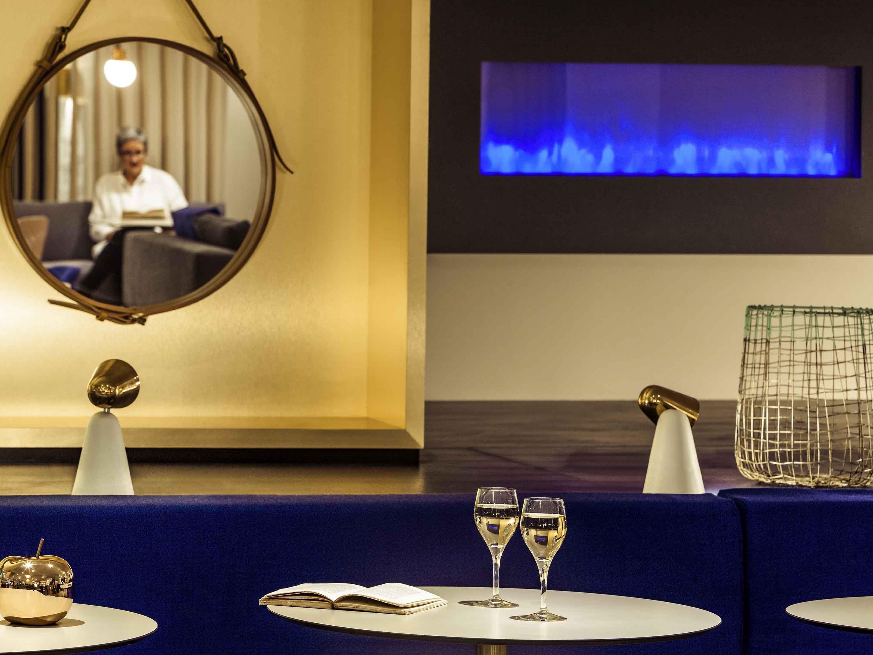 Um saguão de hotel com um espelho redondo, uma tela de televisão, um sofá azul, dois copos de vinho e uma tigela decorativa.