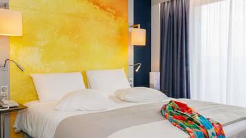 A hotel room with a large bed, white bedding, a colorful throw blanket, a nightstand with a phone, and a wall with a yellow and blue gradient.