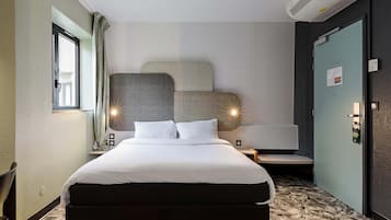 A hotel room with a bed, a desk, a chair, a window, an air conditioning unit, and a door.