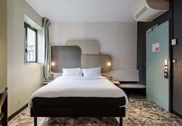 A hotel room with a bed, a desk, a chair, a window, an air conditioning unit, and a door.