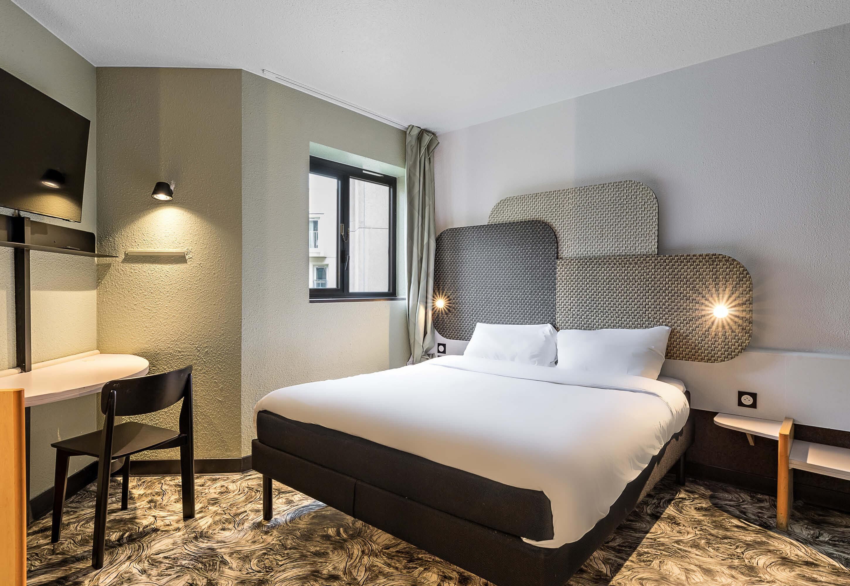 A hotel room with a bed, a desk, a chair, a window, and a wall-mounted TV.