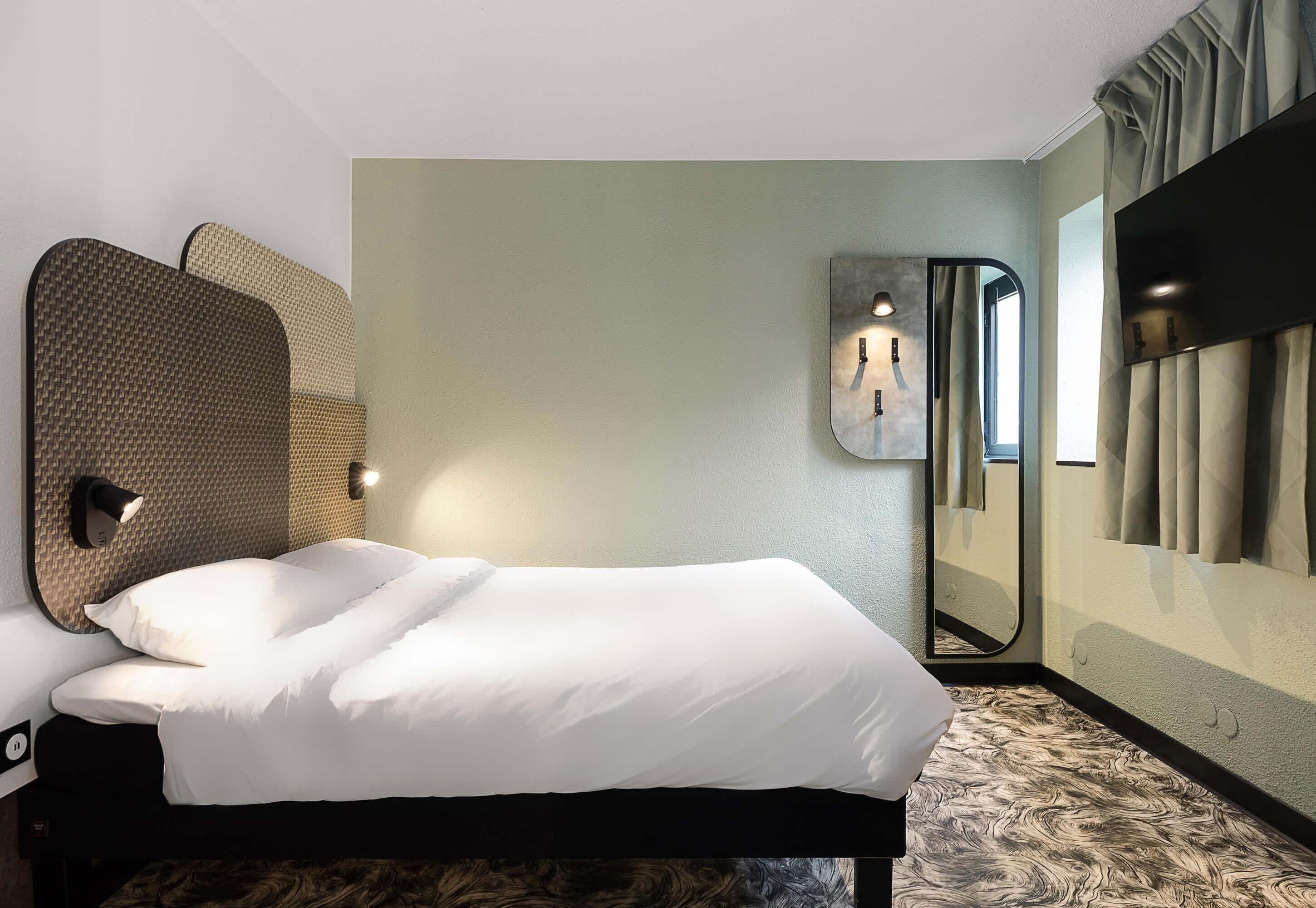 A hotel room with a bed, a wall-mounted light, a mirror, and a large window with curtains.