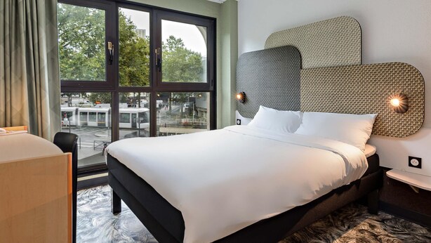 Une chambre d’hôtel avec un grand lit, une vue sur l’extérieur, une tête de lit moderne et une petite table de chevet.