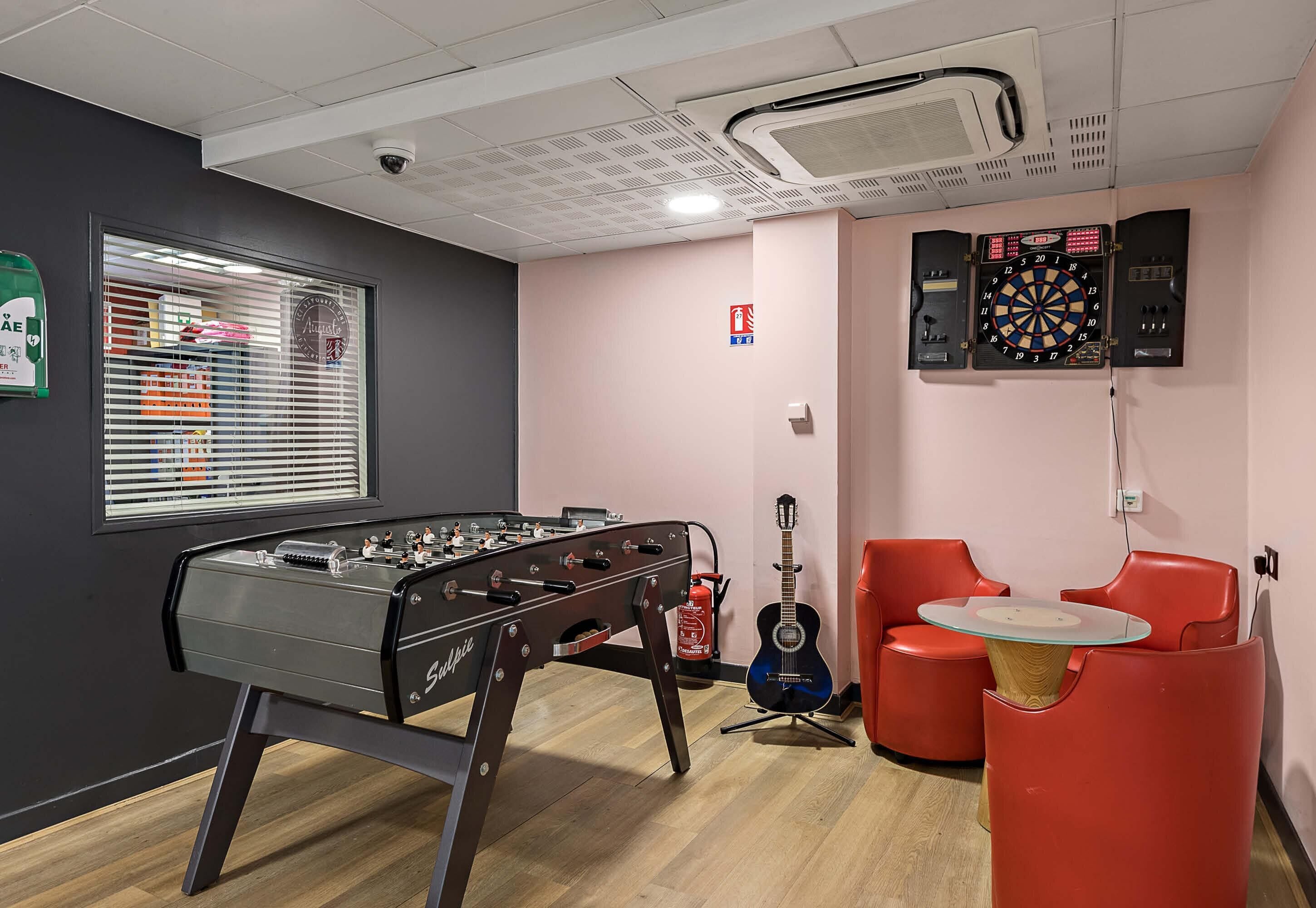 A room with a foosball table, a dartboard, a guitar, and red chairs.