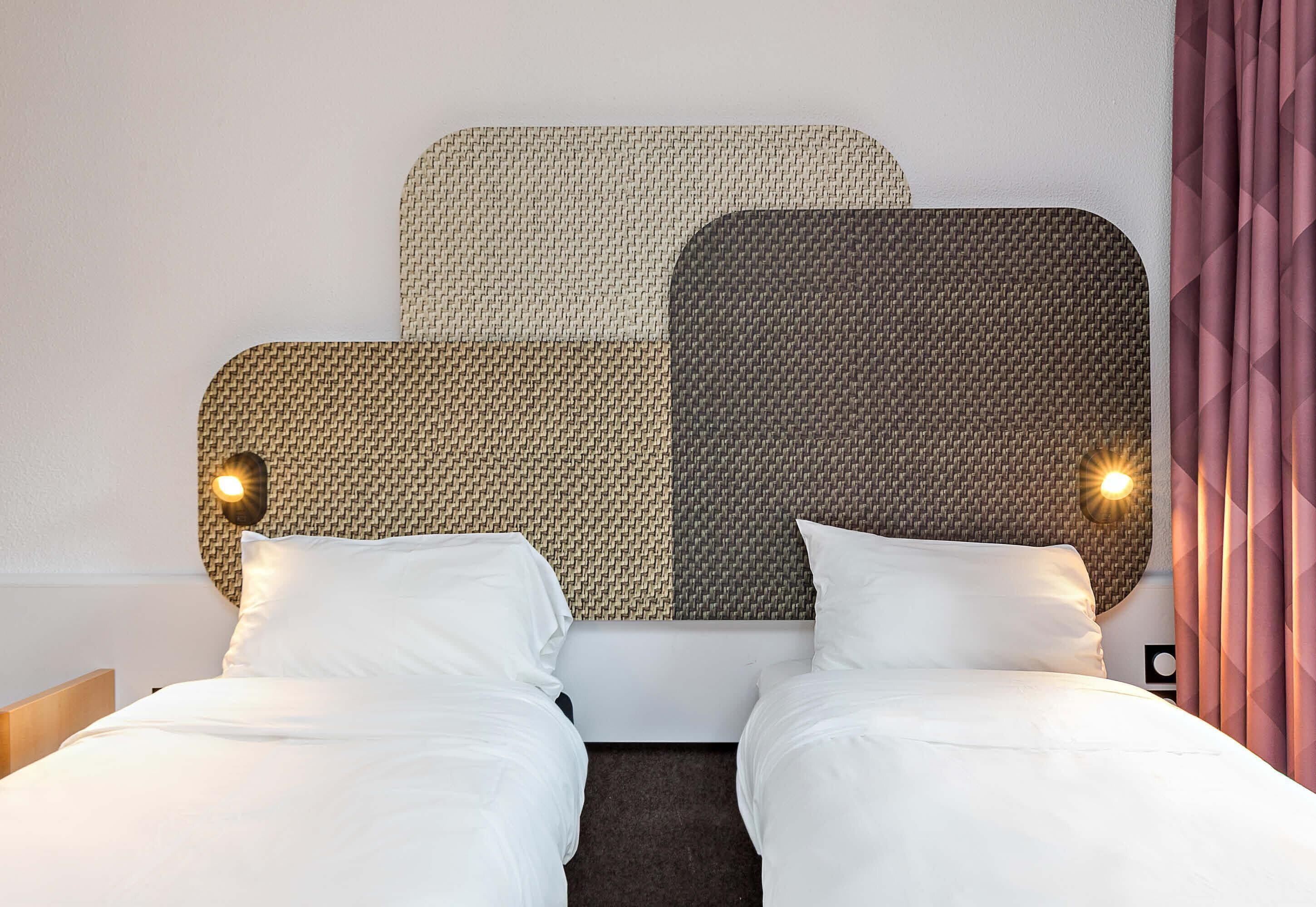 Quarto de hotel com duas camas, cabeceira com padrão trançado e luminária fixada na parede.
