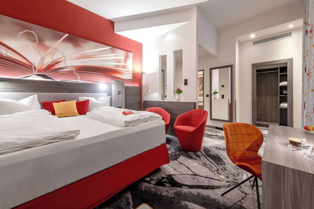 Habitación de hotel moderna con una cama grande, ropa de cama roja y blanca, una silla roja y un escritorio con un florero y fruta.