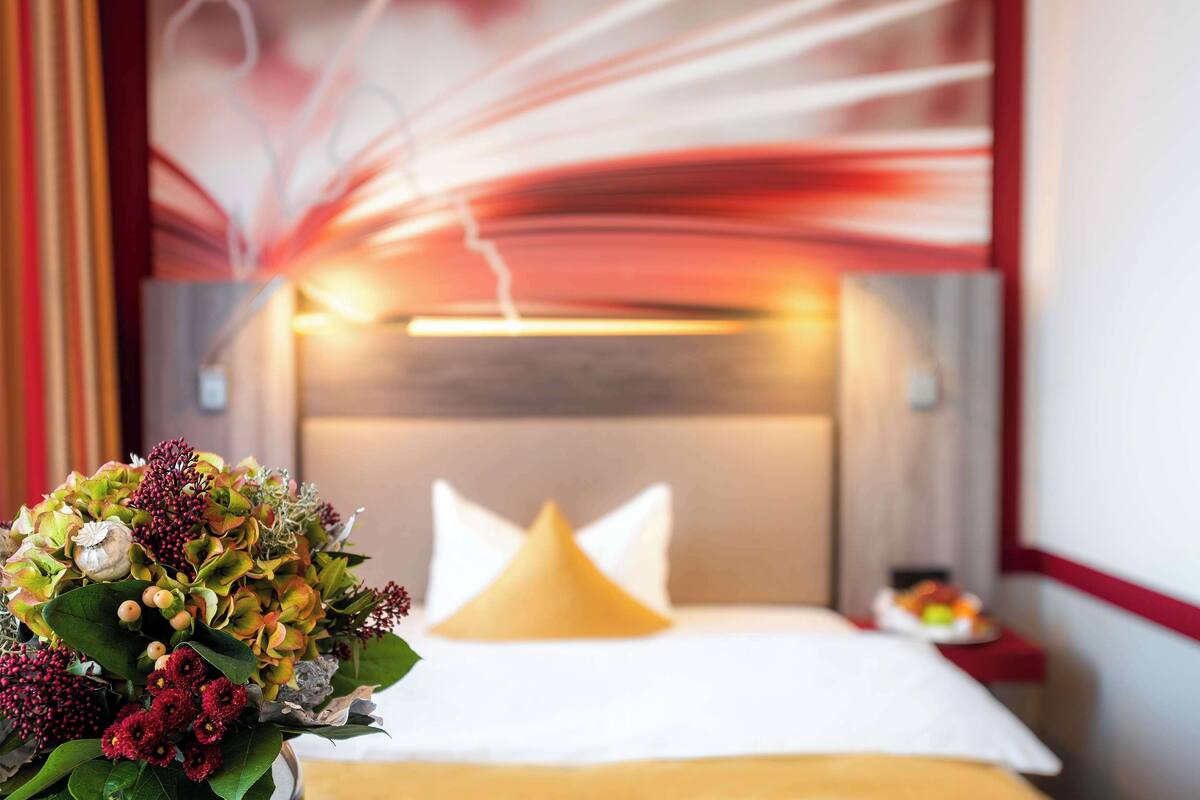 Habitación de hotel con cama, mesita de noche con una bandeja de fruta y un arreglo floral decorativo.