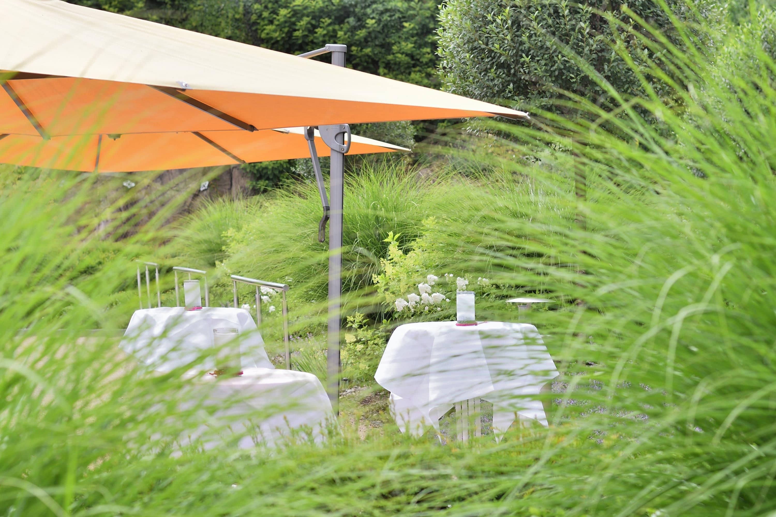 An outdoor dining area with tables covered in white tablecloths, an orange umbrella, and greenery in the background.