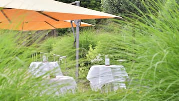 An outdoor dining area with tables covered in white tablecloths, an orange umbrella, and greenery in the background.