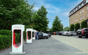 Zona d'aparcament amb estacions de càrrega per vehicles elèctrics, un edifici i cotxes aparcats.