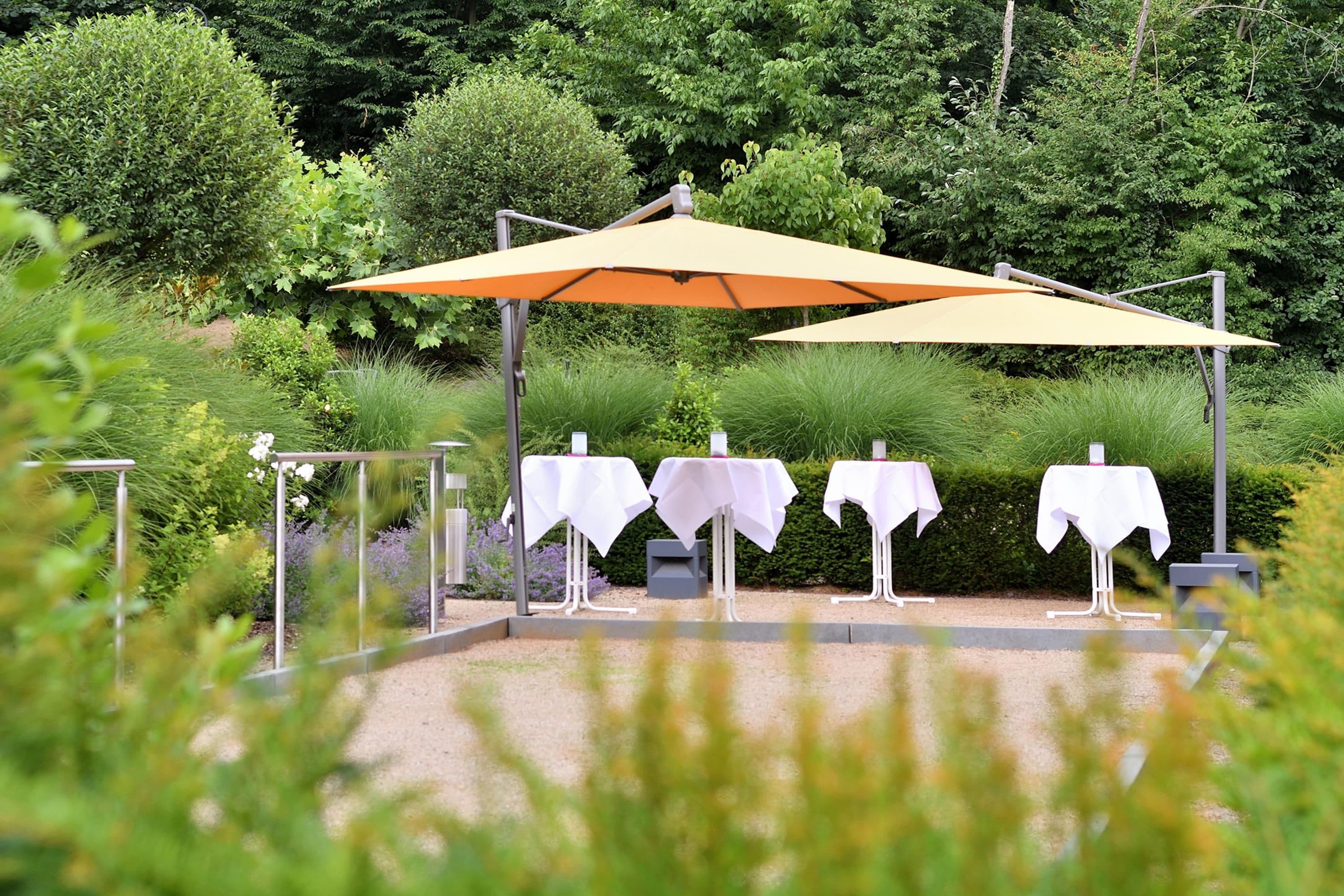 An outdoor seating area with tables and umbrellas, surrounded by greenery.