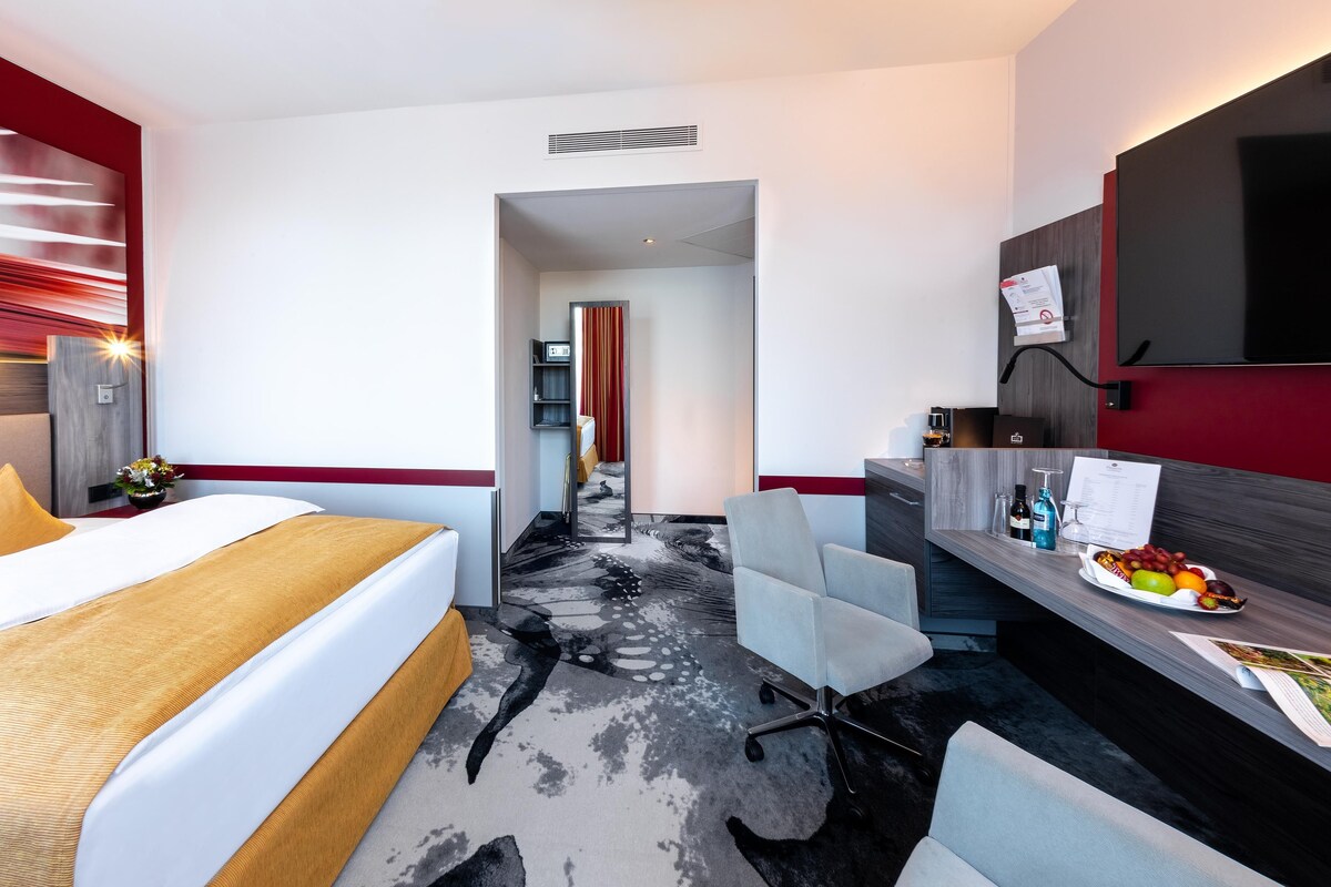 Une chambre d’hôtel moderne équipée d’un lit, d’un bureau avec un ordinateur, d’une chaise et d’une petite table sur laquelle se trouve des fruits.