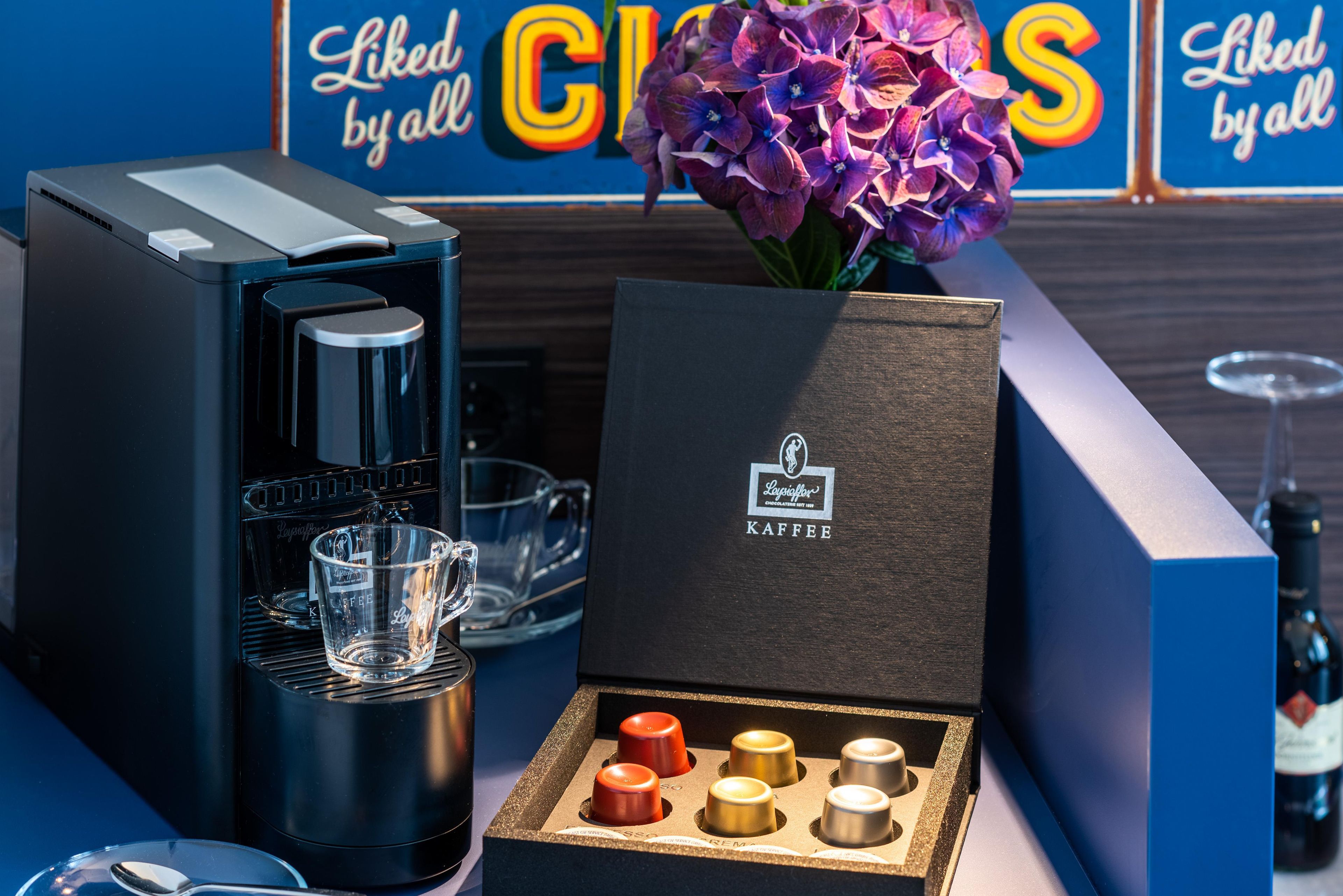 Una cafetera, una taza de vidrio, una caja con una taza de café y un florero con flores.