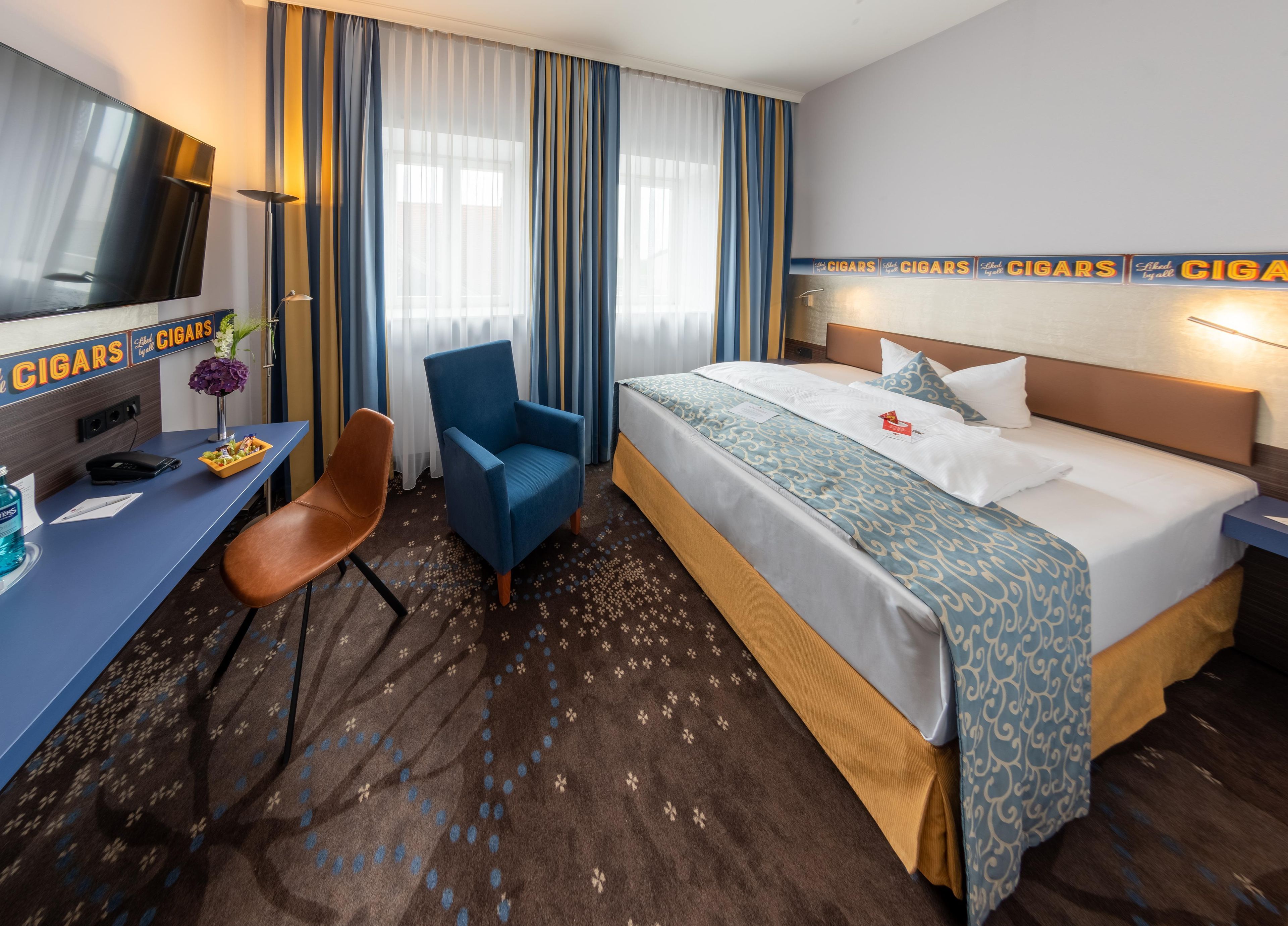 A hotel room with a bed, a desk, a chair, a TV, and a wall with cigar-themed decorations.