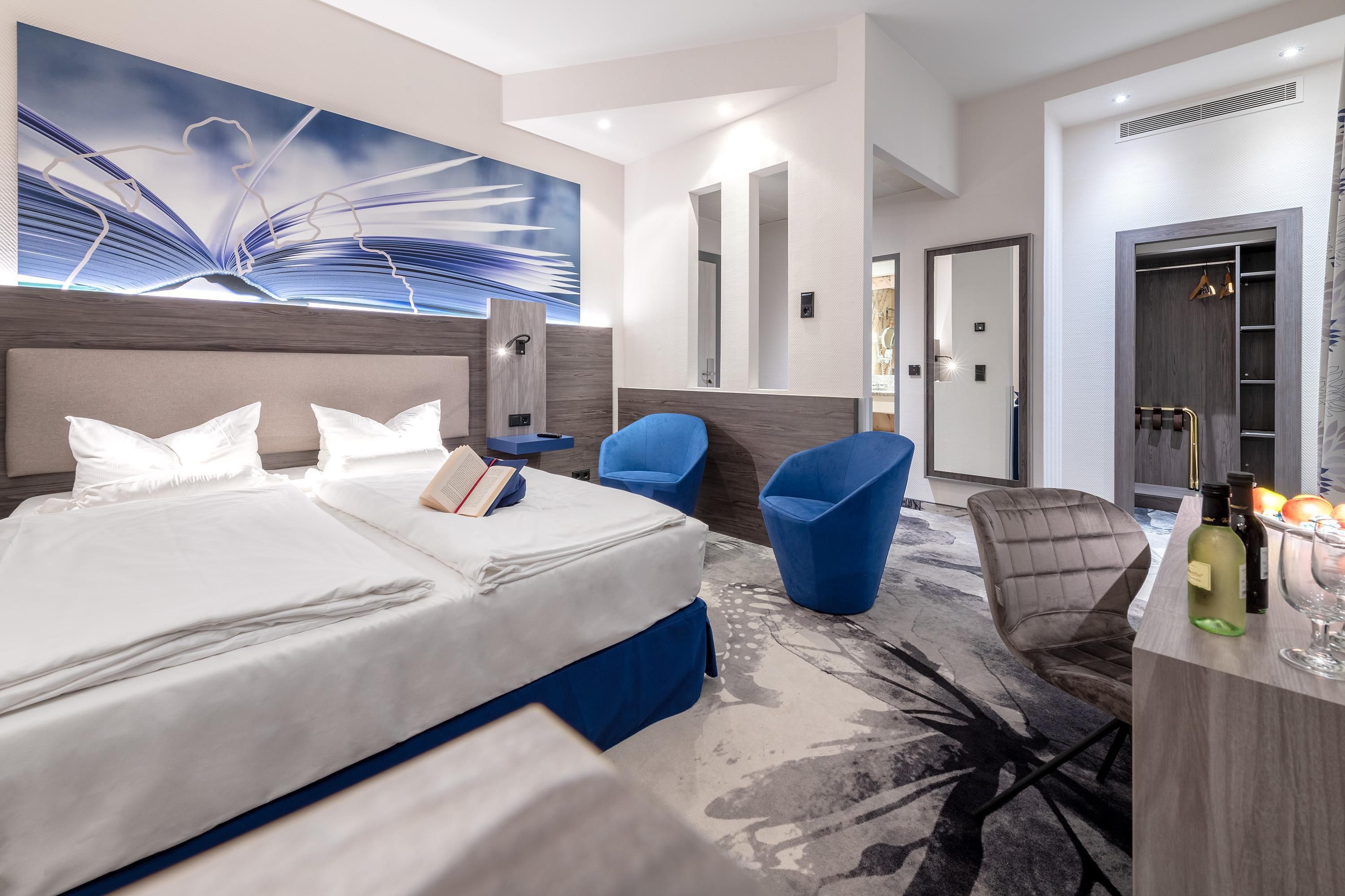 A modern hotel room with a large bed, a seating area with chairs, a vanity with a mirror, and a minibar.
