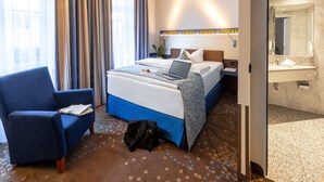 A hotel room with a bed, a desk with a laptop, a chair, a nightstand, a potted plant, and a bathroom with a mirror and sink.