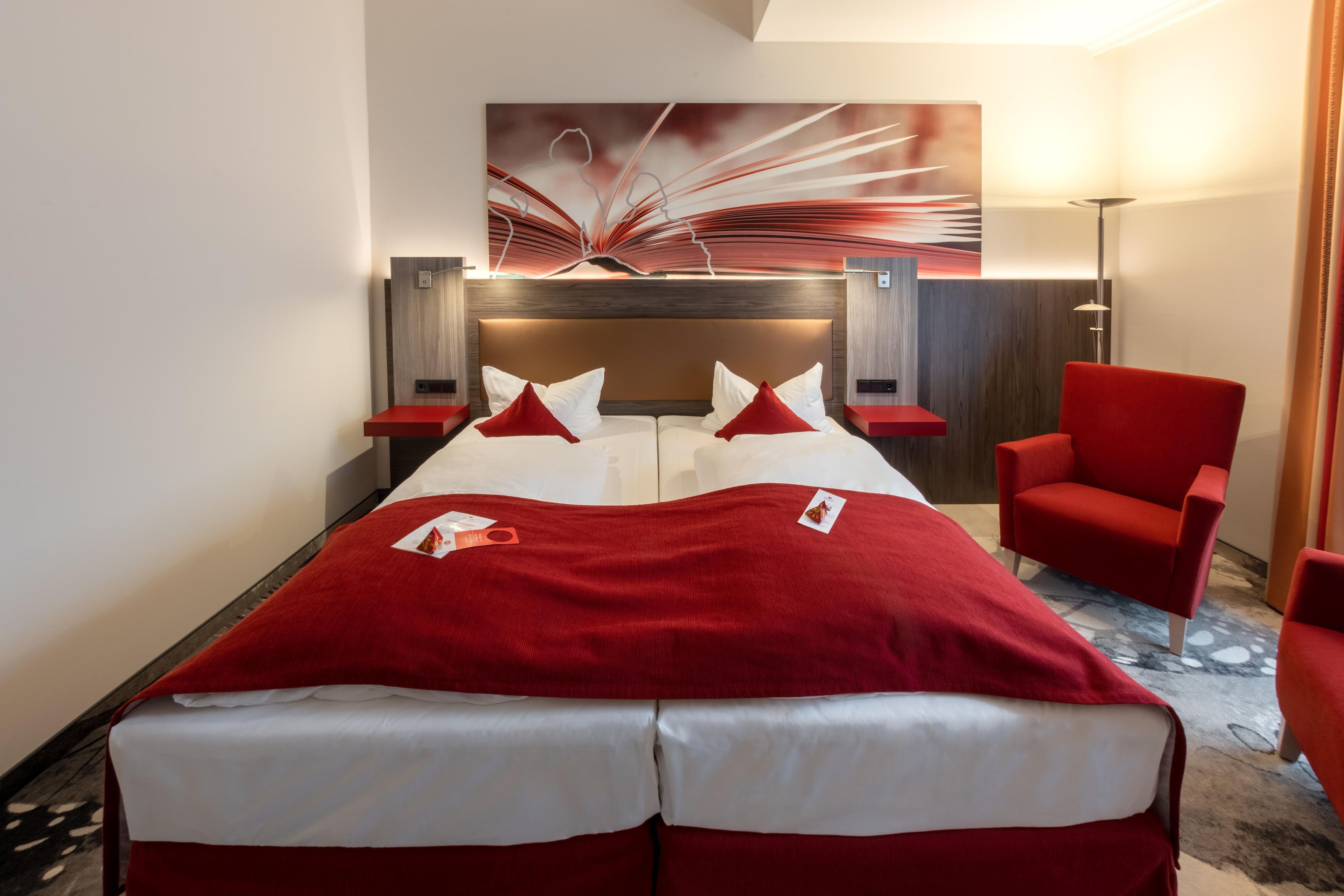 Une chambre d’hôtel avec un lit, des chaises rouges, une couverture rouge et une œuvre d’art murale.