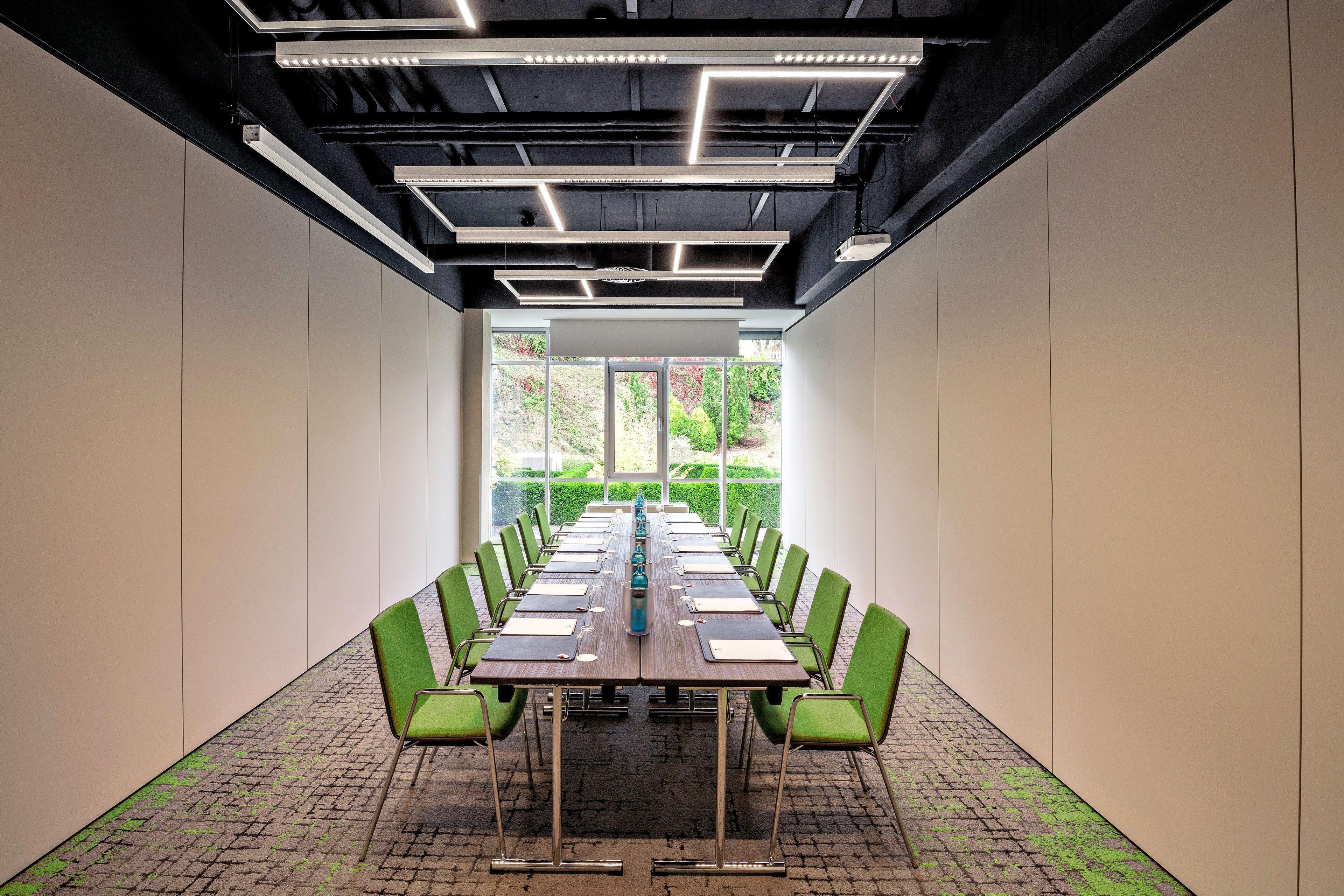 A conference room with a long table, green chairs, and a view of greenery outside.