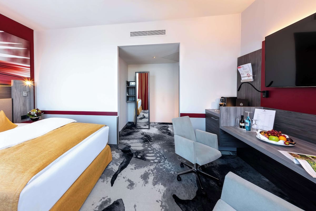 Una habitación de hotel moderna con una cama, un escritorio, una silla, un televisor y un pequeño rincón comedor con fruta.