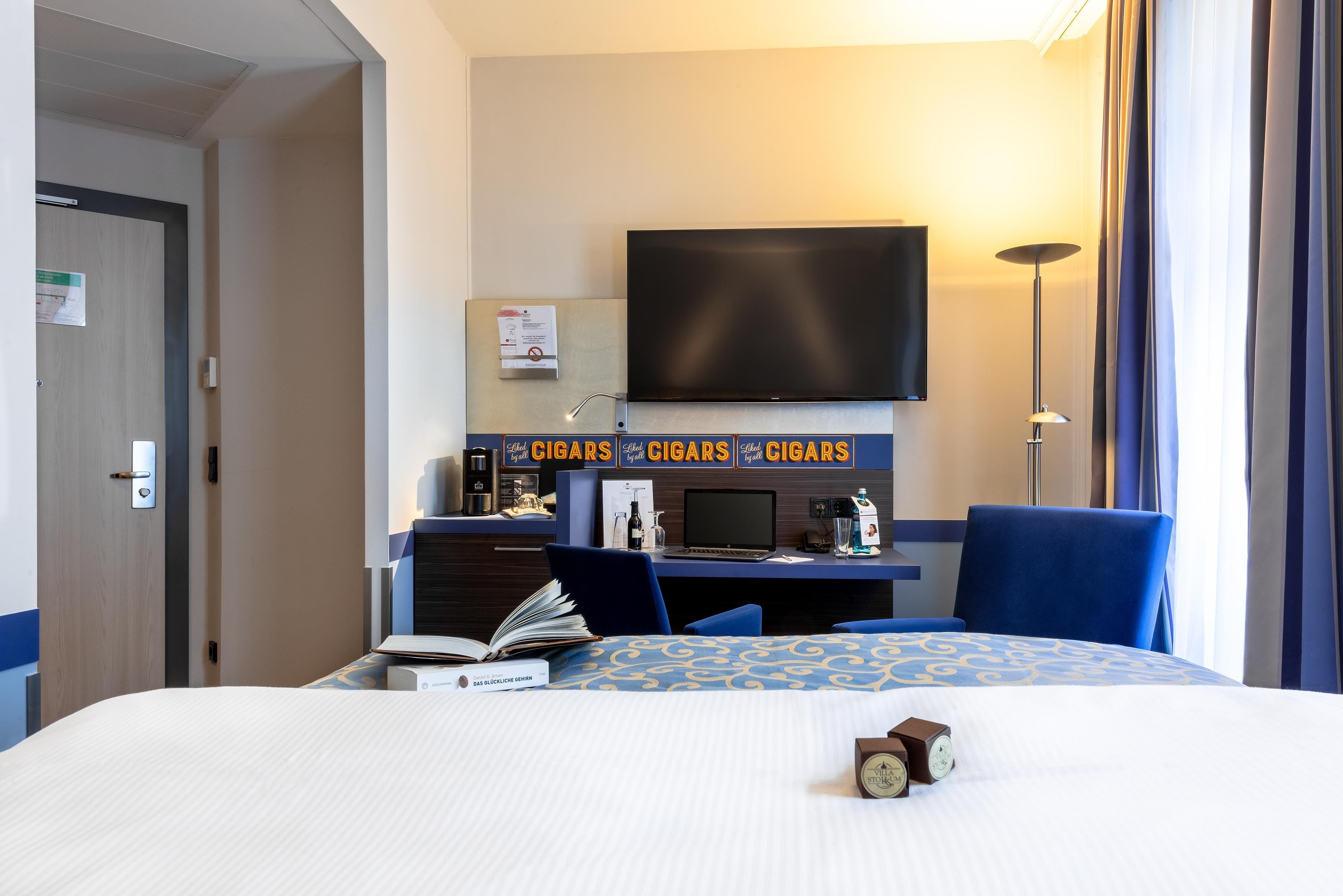 A hotel room with a bed, a desk with a computer, a flat-screen TV, and a window with curtains.