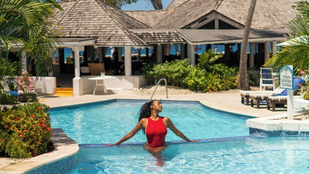 A woman in a red swimsuit standing in a pool with a tropical resort background including palm trees and a thatched-roof building.