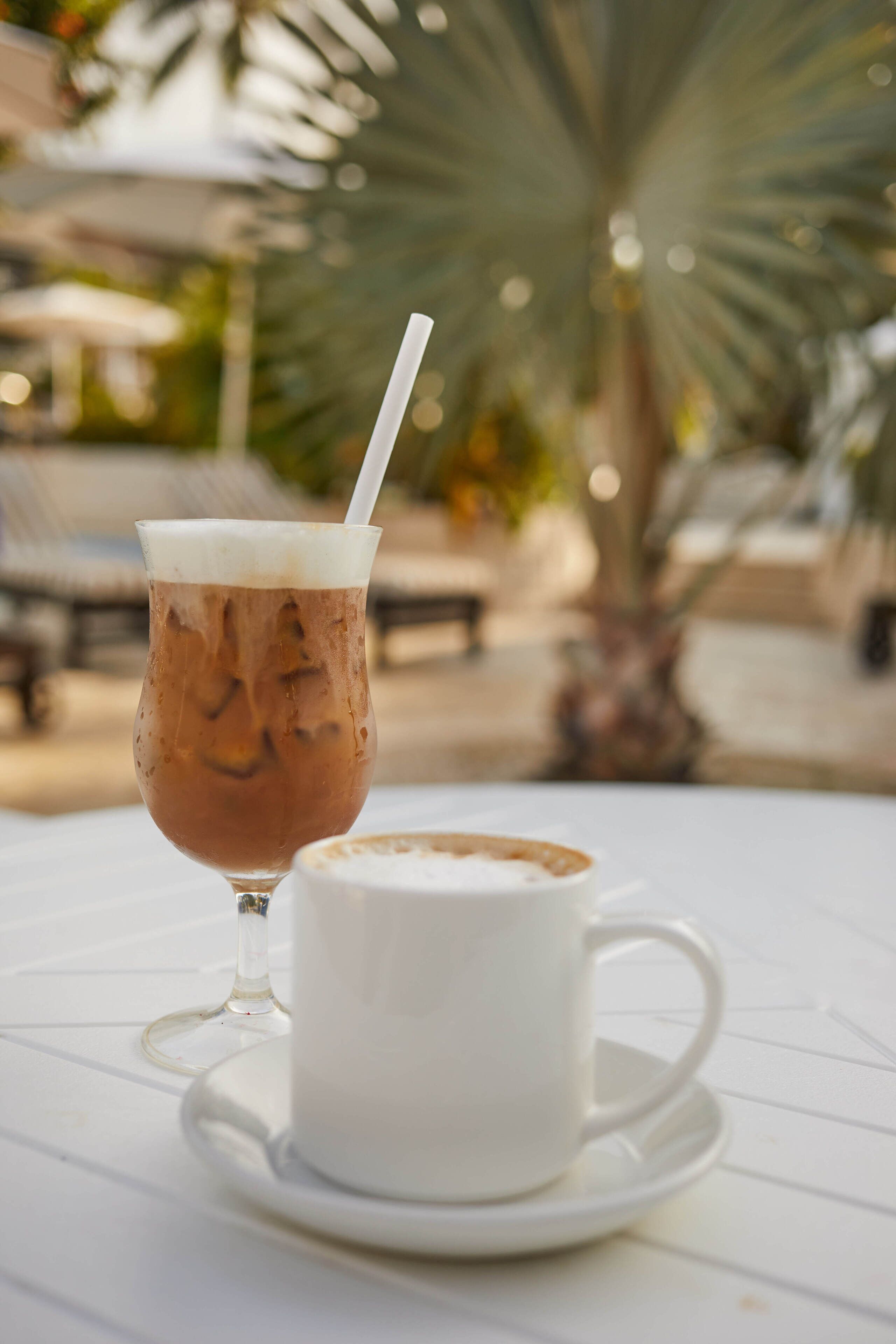 A glass of iced coffee with a straw on a white table, next to a white cup and saucer.