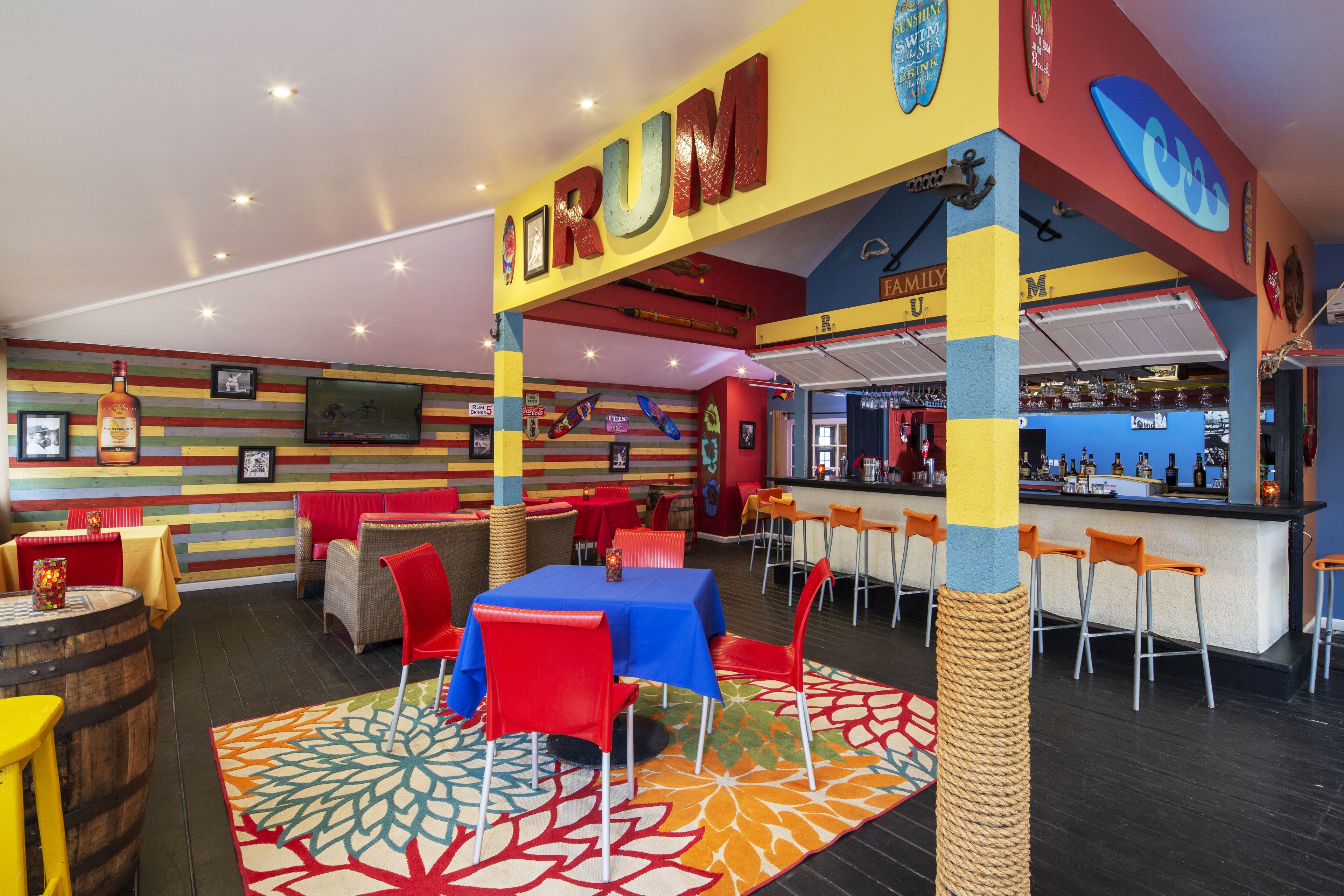 Un espace bar aux couleurs vives, agrémenté d’une enseigne « TRUM », de tables recouvertes de nappes rouges et bleues, et d’un comptoir avec des tabourets.