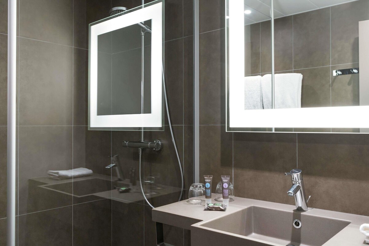 A modern bathroom with a large mirror, a shower, and a sink with toiletries.